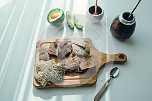 Layout of linen breads served on wooden board with fresh avocado, sauser and calabash in the background, all on white surface with photo