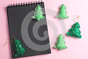 Layout for Merry Christmas to-do list. black notebook on pink background. toys made of felt in the form of green fir trees, flat