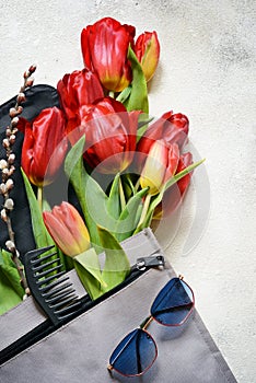 Layout of tulips, backpack and women`s personal belongings on a light-colored concrete background.Flowers, bag, nail polish