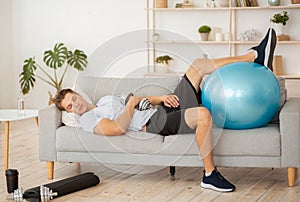 Laziness and workout. Tired guy sleeping on couch with dumbbell and fitness ball photo