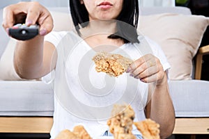 Lazy Asian woman eating fried chicken hand holding remote tv, unhealthy lifestyle concept