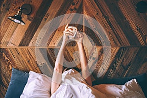 Lazy female lying under the white blanket on the  linen bed and holding the fresh coffee cup in the early morning. Lazy day off