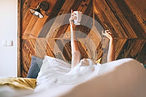 Lazy female lying under the white blanket on the  linen bed and holding the fresh coffee cup in the early morning. Lazy day off