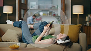 Lazy girl enjoying headset music laying couch. Carefree lady browsing smartphone