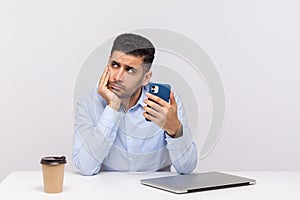 Lazy inefficient unproductive man employee sitting in office workplace, holding smartphone and looking away