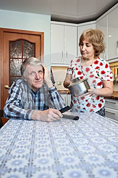 Lazy mature husban lounges in the kitchen, his wife makes help on housework