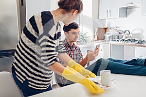 Lazy man looking at phone while his angry girlfriend cleaning his dishes in kitchen and talking to him. Family arguement photo