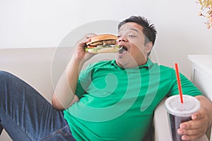 Lazy overweight man eating hamburger while laying on a couch