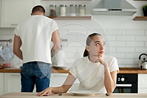 Lazy thoughtful wife sitting at kitchen table while husband cook