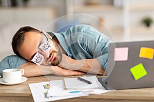 Lazy unproductive young guy wearing funny sticky notes with open eyes on his glasses, sleeping at workplace