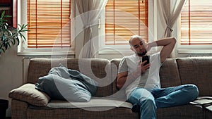 Lazy weekend. Boring bald bearded caucasian man sitting on sofa and using smartphone. Tracking right shot. Concept of