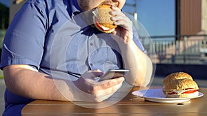 Lazy young man eating unhealthy burger and scrolling smartphone application