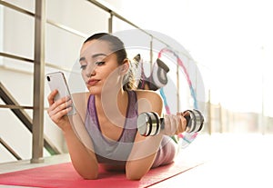 Lazy young woman with dumbbell and smartphone on yoga mat