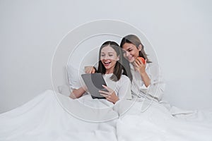 LBGT Couple of cute lesbian women waking up in the morning on white bed in bedroom while laughing and looking at tablet