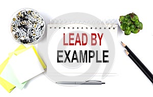 LEAD BY EXAMPLE text on notebook with office supplies on white background