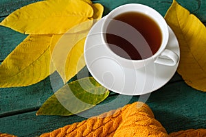 Leaf fall mood, white cup of coffee with a knitted scarf and fallen leaves on wooden background