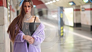 Leaned on the wall model girl in synthetic fur coat bared her shoulders waiting in metro or subway platform in slow
