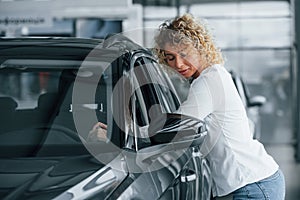 Leaning on the car. Woman with curly blonde hair is in autosalon