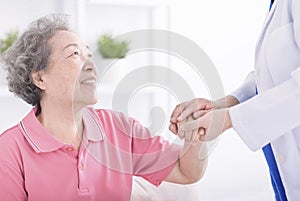 leaning forward to smiling elderly holding her hand in palms. Woman caretaker in white coat supporting encouraging old person