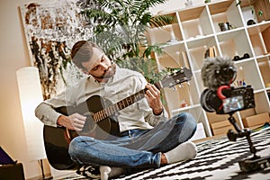 Learning guitar. Bearded male music blogger sitting on the floor at home and recording new guitar lesson for Youtube