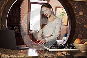 Learning by the videos. Pretty young woman standing in the modern kitchen near gas stove and preparing food using laptop