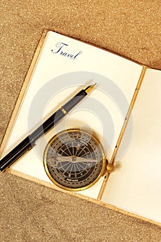 Leather journal, with the word travel on a page, with a fountain pen and antique compass in sand.