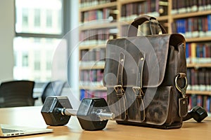 leather satchel on library table, dumbbells handle sticks out, laptop adjacent