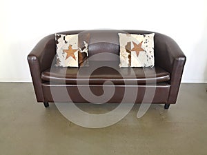 Leather sofa, brown,  with two decorative pillows on a beige-brown floor in front of a white wall.