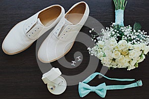 Leather white groom wedding shoes on the wooden background in sun rays. Rings, bow tie and cufflinks with elegant bouquet. Elegant