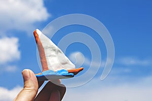 On the left, fingers are holding a brown boat sculpted from plasticine against a blue sky with white clouds