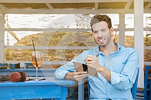 Left-handed guy sitting in a beach bar drinking cocktail and writing as freelancer