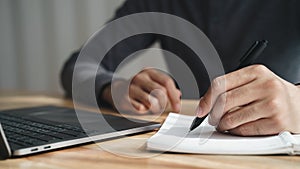 Left handed man writes in a notebook on the table with laptop computer