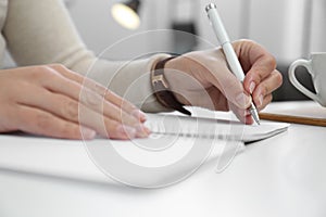 Left-handed woman writing in notebook at table indoors, closeup