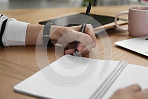 Left-handed woman writing in notebook at wooden table, closeup