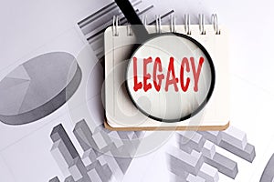 Legacy word highlighted with magnifying glass on notepad over financial graphs and charts