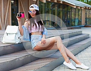 Leggy young cheerful hipster woman sitting on the steps in city wearing vintage pink top, jeans shorts and American flag sunglasse