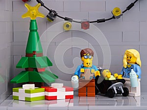 A Lego family - father and mother with two babies minifigures standing near a Christmas tree that has presents under it