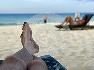 Legs on a beachchair and blurry man reading a book on the beach