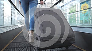 Legs of business woman going through hall of terminal with her luggage. Young girl in high heels walking on escalator