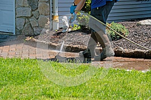 Legs and feet wearing rubber boots and hands holding a power washer wand spraying soapy water on a brick walkway sidewalk