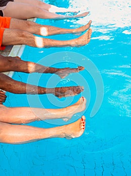 Legs of four friends common people different race sitting by the swimming pool in sunny day.