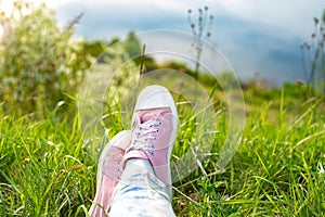 Legs of the girl in jeans and white and pink on the green grass in the summer near the river
