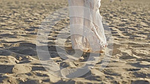 Legs of girl wearing white long dress walking barefoot sand on sea beach or desert. Slow motion. Woman walks to camera