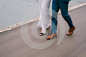 Legs of the groom in blue trousers and the bride in a white dress are walking along the asphalt road over the sea. Close