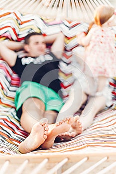 Legs of a happy stylish family playing with cute daughter, relaxing in hammock on summer vacation in evening sun light on the