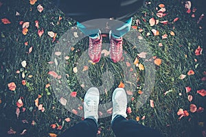 Legs of a man and a girl in sneakers on grass, strewn with autumn leaves, top view