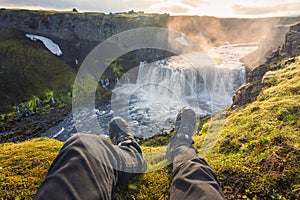 Legs of man sitting on edge of mountain with Axlafoss waterfall flowing in canyon