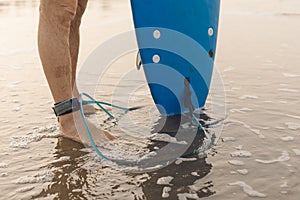 Legs of man standing on sandy seaside with leash attached to ankle, holding surfboard. Athlete preparing for surfing.