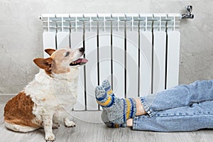 Legs on a radiator . A woman in warm knitted woolen socks and cute corgi dog near a home heater in the cold autumn-winter season.