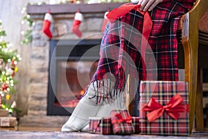 Legs in a socks of disabled women sitting on armchair covered plaid near fireplace and christmas tree  and pakking gift boxes